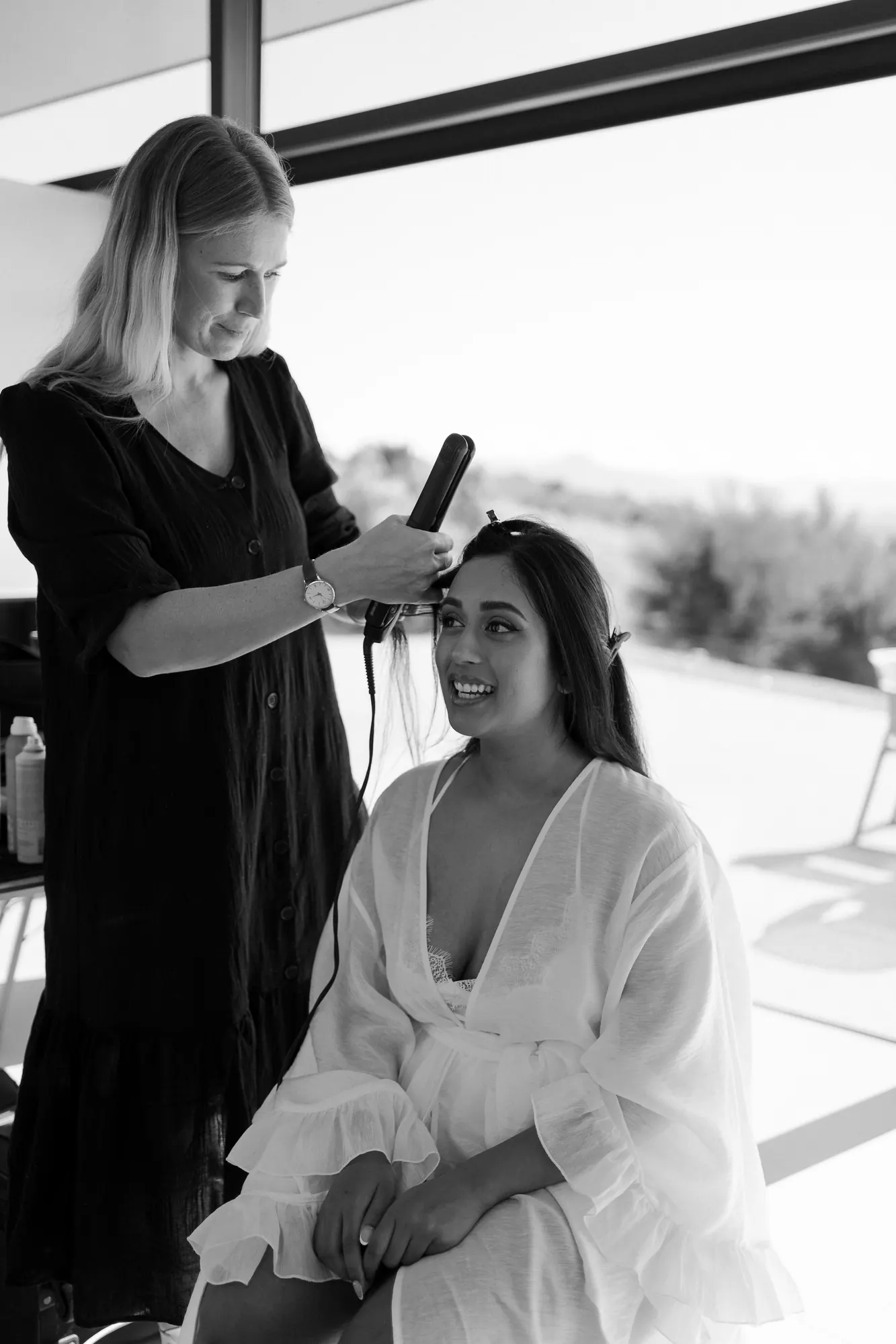Stacey styling a smiling bride's hair with a flat iron, in black and white